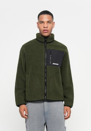 Fuzzy olive green fleece jacket with a high collar, zip closure, and a black chest pocket featuring a logo. Textured fabric and relaxed fit.