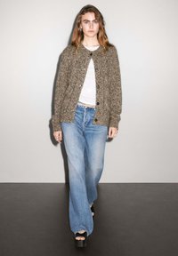 Brun stickad cardigan med knappar fram, bärs över en vit topp. Blå vida jeans och svarta platåslip-ons fulländar outfiten.