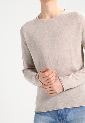 Personne portant un pull beige à manches longues et un jean bleu, les mains jointes au niveau de la taille sur un fond clair uni.