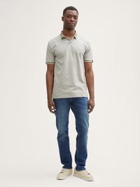 Camisa polo caqui con cuello y ribete en contraste, combinada con jeans azules y zapatillas de color claro.