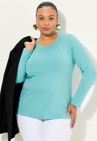 Türkises Langarmshirt aus einem weichen Stoff mit Rundhalsausschnitt, kombiniert mit einem schwarzen Blazer, der über der Schulter getragen wird. Weiße Hose sichtbar.