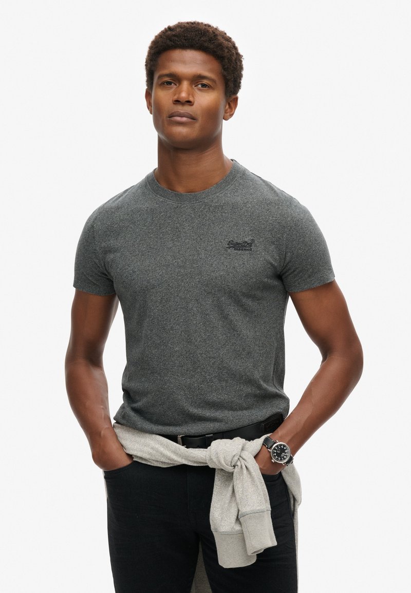 Grå kortærmet T-shirt med rund hals og lille logo, parret med sorte jeans og en lysegrå sweater bundet om taljen.