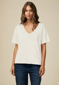 Bílý T-shirt s V-výstřihem a krátkými rukávy, vyrobený z měkké tkaniny. Volný střih s mírně zakřiveným lemem, kombinovaný s tmavě modrými džíny.