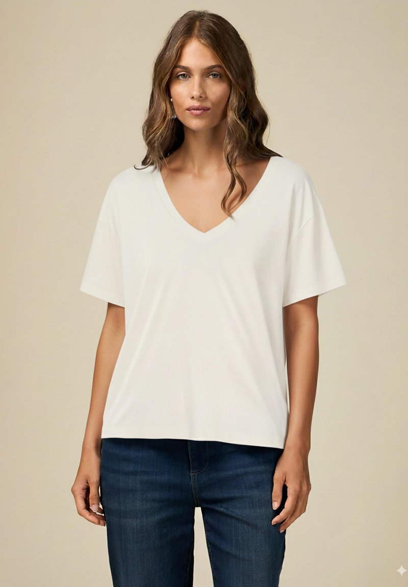 Bílý T-shirt s V-výstřihem a krátkými rukávy, vyrobený z měkké tkaniny. Volný střih s mírně zakřiveným lemem, kombinovaný s tmavě modrými džíny.