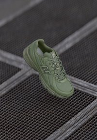 Olíva zöld sneaker sima, texturált felső résszel, párnázott bokarésszel és fűzőkkel; az oldalán fényvisszaverő logóval, egy rácsos felületen elhelyezve.
