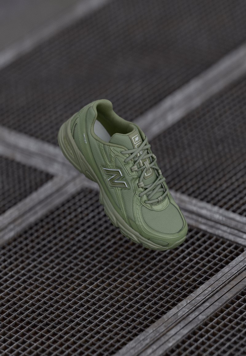 Olíva zöld sneaker sima, texturált felső résszel, párnázott bokarésszel és fűzőkkel; az oldalán fényvisszaverő logóval, egy rácsos felületen elhelyezve.