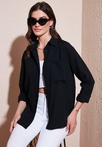 Black button-up shirt with vertical ribbed texture, single chest pocket, worn over a white crop top, paired with white jeans and black sunglasses.