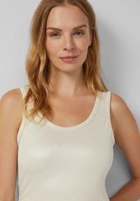 Vrouw met golvend blond haar en blauwe ogen, gekleed in een wit geribbeld tanktop, staand tegen een effen lichtgrijze achtergrond.