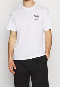 Valkoinen puuvillapaita lyhyillä hihoilla ja pyöreällä kaula-aukolla, jossa on musta graafinen muotoilu rinnassa ja suora helma.