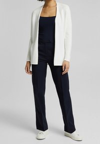 Bílý kardigan, námořnický top a námořnické kalhoty. Kardigan má otevřený přední design, hladkou strukturu a dlouhé rukávy. Tenisky jsou bílé.