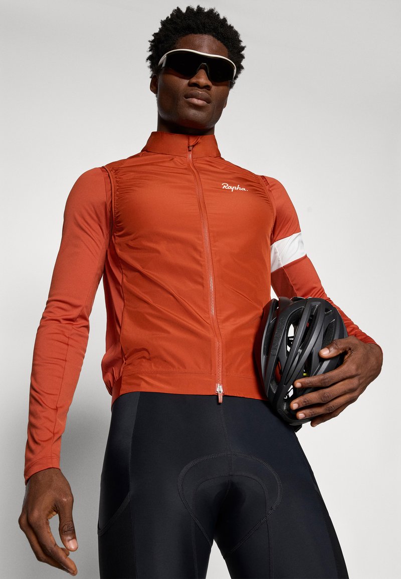 Rote, leichte Radjacke mit hohem Kragen, Frontreißverschluss und weißen Akzenten. Schwarze gepolsterte Radsportshorts und Helm in der Hand.