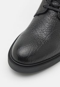 Zapato de cuero negro con una superficie texturizada, punta redondeada y suela de goma negra. El detalle de cordones en la parte superior agrega funcionalidad.