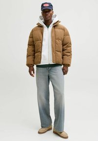 Brūns pūkains jaka ar kordūras faktūru, pelēka kapuce zem tās, gaiši zili džinsi, bēši apavi un tumši zils cepure ar sarkanu logotipu.