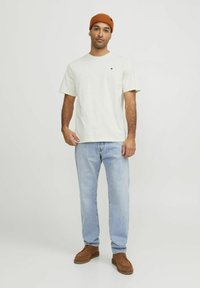 Bomull t-shirt i ljusgrädde med korta ärmar och en liten röd logotyp, tillsammans med ljusblå jeans och bruna mockaskor.