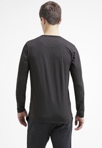 Muž v jednoduchém černém tričku s dlouhým rukávem a tmavých kalhotách, stojící zády k bílému pozadí.