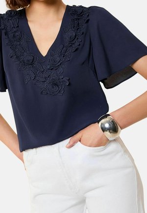 Granatowa bluzka z kwiatowym koronkowym dekoltem w kształcie litery V, krótkie falbaniaste rękawy, noszona z białymi spodniami i szeroką srebrną bransoletą na rękę.