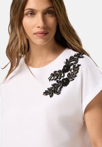Camiseta de algodón blanca con intrincado bordado floral negro en el hombro izquierdo, adornada con cuentas texturizadas y lentejuelas para mayor detalle.