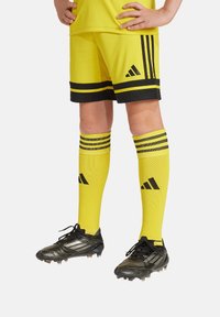 Uniforme de sport jaune avec un t-shirt à col ras du cou et un short assorti à rayures noires. Chaussures de football noires et chaussettes montantes à rayures.