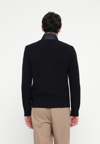 Homme debout, de dos, portant un pull en tricot noir sur une chemise noire et un pantalon beige, sur fond blanc uni.