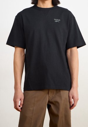 Man draagt een effen zwart Reebok T-shirt met korte mouwen en een bruine broek, staand tegen een neutrale achtergrond.