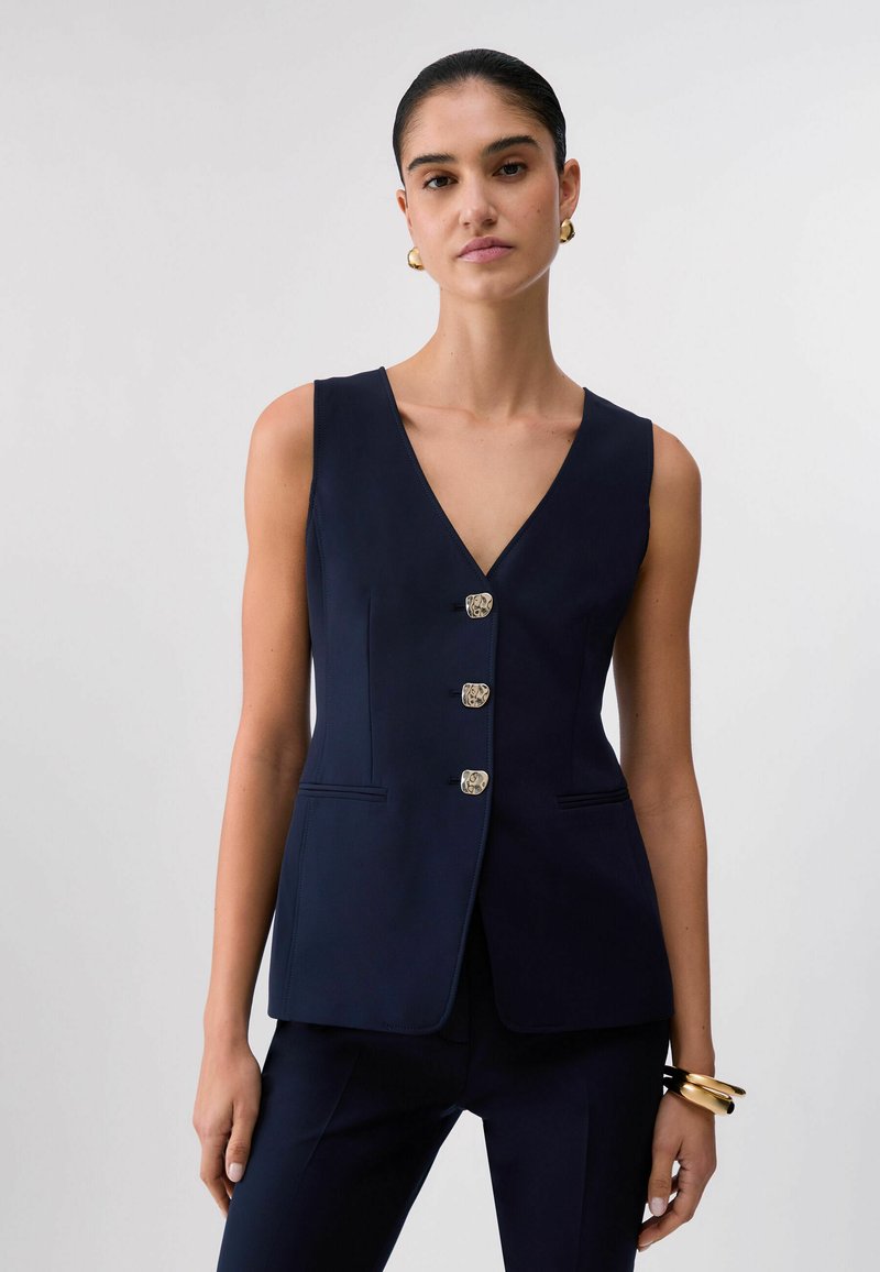 Kvinde iført en ærmeløs navy vest med tre dekorative knapper, suppleret med matchende navy bukser og guld øreringe i hulringe.