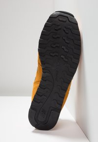 Semelle en caoutchouc noir avec un motif de profil en zigzag, attachée à un dessus en suède jaune. Présente le logo "New Balance" imprimé sur le côté.