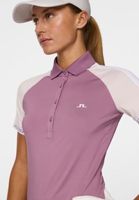Polo shirt viola e crema con colletto, dettagli con bottoni e logo sul petto. Presenta maniche corte e una texture liscia.