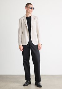 Blazer beige texture con due tasche sopra una maglietta nera, abbinato a pantaloni neri e scarpe nere, su uno sfondo neutro.