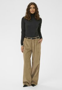 Mujer con cabello rizado, vistiendo un suéter de cuello alto gris oscuro y pantalones de pana anchos color beige con un cinturón negro, de pie frente a un fondo blanco.