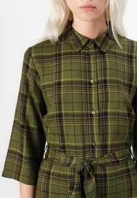 Green plaid shirt dress with a collar, three-quarter sleeves, button front, and a tied waist detail. Textured fabric with contrasting patterns.