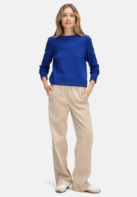 Kék bordázott kötött pulóver kerek nyakkivágással és hosszú ujjal, bézs, bő szárú kordbársony nadrággal és fehér cipőkkel párosítva.