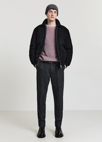 Schwarze Sherpa-Jacke mit Reißverschluss über einem hellrosa gerippten Pullover, graue Hosen mit strukturiertem Muster und schwarze Stiefeletten, dazu eine graue Strickmütze.