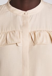 Blouse beige avec un col rond, une fermeture à boutons et des détails froncés sur le devant. Tissu lisse et léger avec un léger éclat.