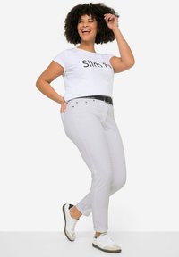 T-shirt blanc arborant le texte "Coupe ajustée", associé à un pantalon ajusté gris clair, une ceinture noire et des baskets blanches décontractées avec des accents foncés.
