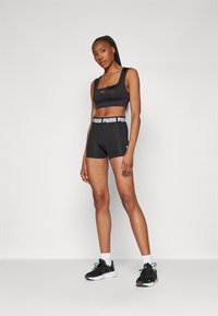 Conjunto de sujetador deportivo negro y pantalones cortos con acentos de logotipo. El sujetador tiene un escote cuadrado; los pantalones cortos tienen una cinturilla elástica amplia. Zapatillas deportivas negras y calcetines blancos.