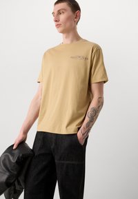 Beige bomulls-t-shirt med korta ärmar, med en liten tryckt logotyp i svart nära axeln, parat med mörka denimbyxor och en svart jacka.