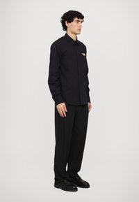 Black button-up shirt with a collar, long sleeves, and a front pocket. Worn with black trousers and shoes, showcasing a minimalist design.