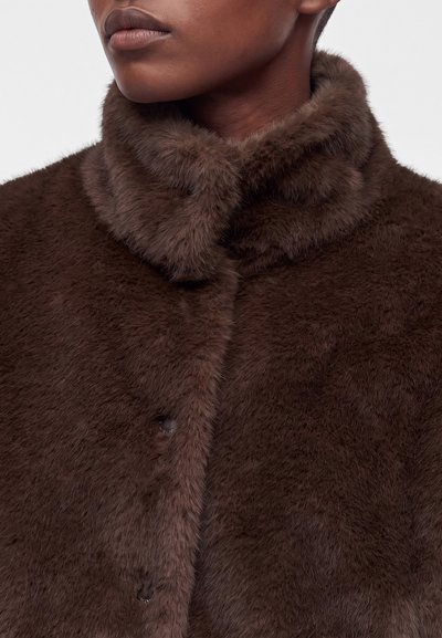 Gros plan d'une personne portant un manteau en fausse fourrure douce de couleur brun foncé avec un col montant sur un fond neutre.