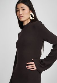 Brązowa dopasowana sukienka z długimi rękawami i falbaniastym dekoltem. Charakteryzuje się prążkowaną teksturą; model nosi duże złote kolczyki.