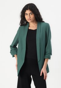 Blazer verde con una vestibilità rilassata, colletto a scialle e maniche arrotolate. Realizzato in tessuto liscio, presenta un design elegante e non strutturato.