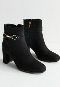 Botas de tobillo de ante negro con punta cuadrada, tacón grueso, cremallera lateral y una tira decorativa de hardware dorado alrededor del tobillo.