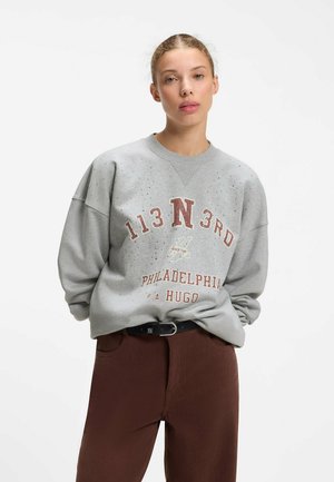 Ung kvinde med lyst hår sat op i en knold iført en grå oversized sweatshirt og brune bukser, stående mod en simpel hvid baggrund.