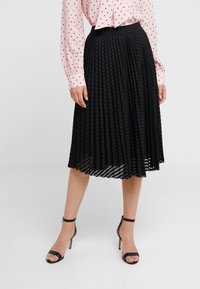 Gonna nera plissettata con finitura testurizzata, dotata di una vita aderente. Abbinata a una blusa rosa chiaro a pois e tacchi neri con cinturini.