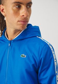 Könnyű anyagból készült kék cipzáras kapucnis pulóver. Bordás textúrával, mellkasi logóval és kontrasztos sárga oldalsávokkal rendelkezik.