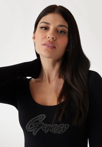 Black long-sleeve top with a scoop neck, featuring a silver rhinestone logo on the front. Smooth texture with a fitted silhouette.