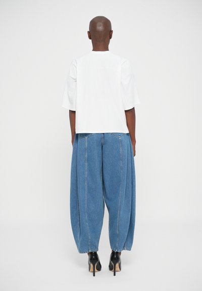 Persoon in een witte korte mouwen top, losse blauwe denim broek en zwarte hoge hakken, staande met de rug naar de camera op een effen achtergrond.
