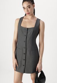 Šedé pruhované šaty s čtvercovým výstřihem, bez rukávů, předními knoflíky a bočními kapsami. Modelka drží černou tašku.