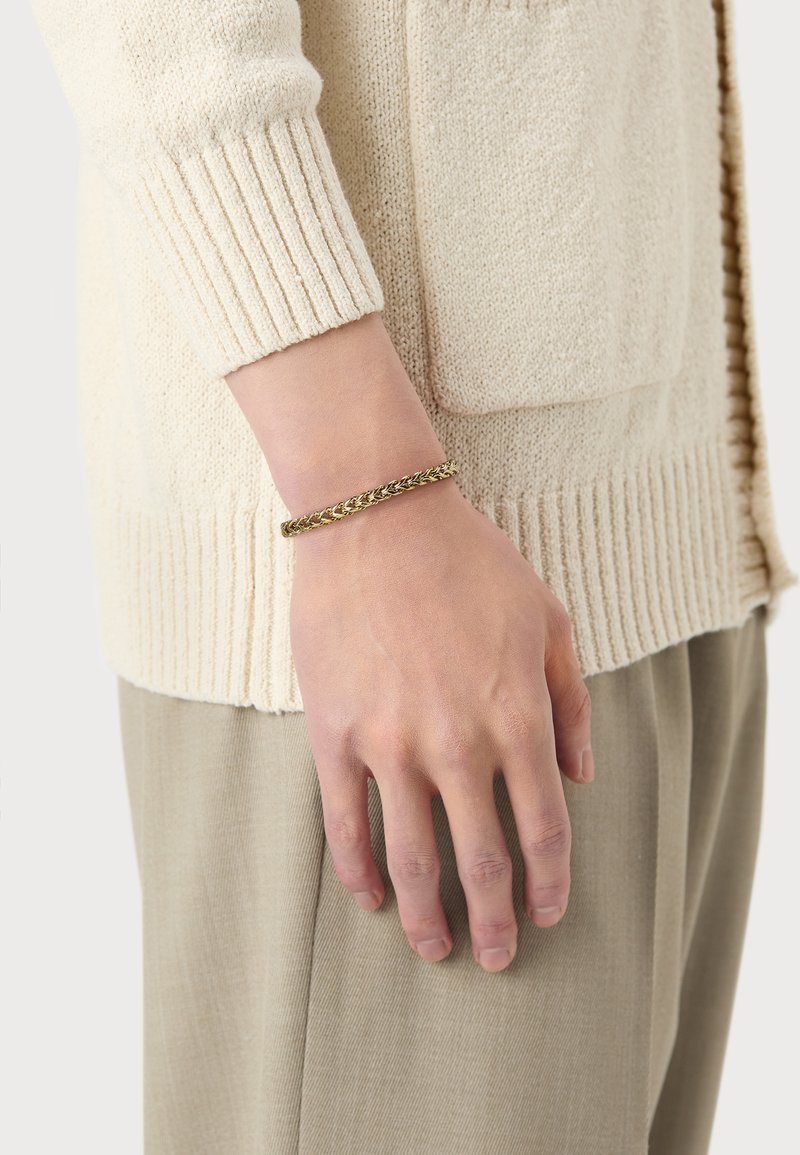 Goldener geflochtener Armreif, der an einer Hand getragen wird, kombiniert mit einer hellbeigen Strickjacke, die gerippte Bündchen und eine frontale Tasche hat.