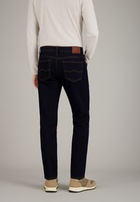 Dunkelblaue Jeans mit orangefarbenen Nähten, fünf Taschen und einem braunen Lederpatch auf der Rückseite; kombiniert mit hellen Schuhen und einem grauen Langarmshirt.