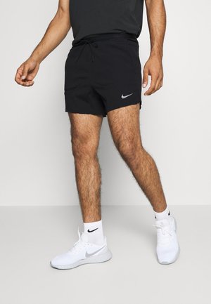 Mężczyzna ubrany w czarne spodenki Nike, białe skarpetki Nike i białe buty do biegania Nike, stojący na jednolitym jasnym tle.
